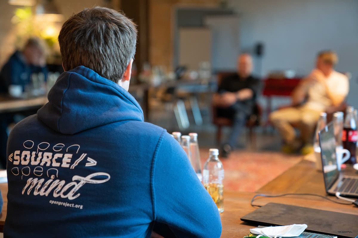 Eine Person trägt ein dunkelblaues Sweatshirt mit Kapuze, auf dessen Rücken das OpenProject-Logo prangt. Das Logo besteht aus stilisiertem Text und einem grafischen Designelement in Weiß.