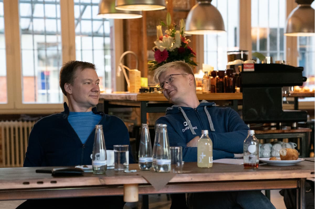 Zwei Männer sitzen an einem hölzernen Esstisch mit Essen und Getränken und führen ein Gespräch.